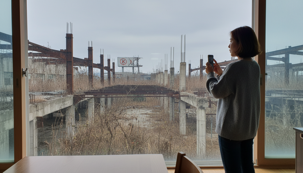 거대한 공장 부지에 잡초가 무성하고 녹슨 철골 구조물만 남아 있는 황량한 풍경, 멀리서 빛바랜 기업 로고가 보임